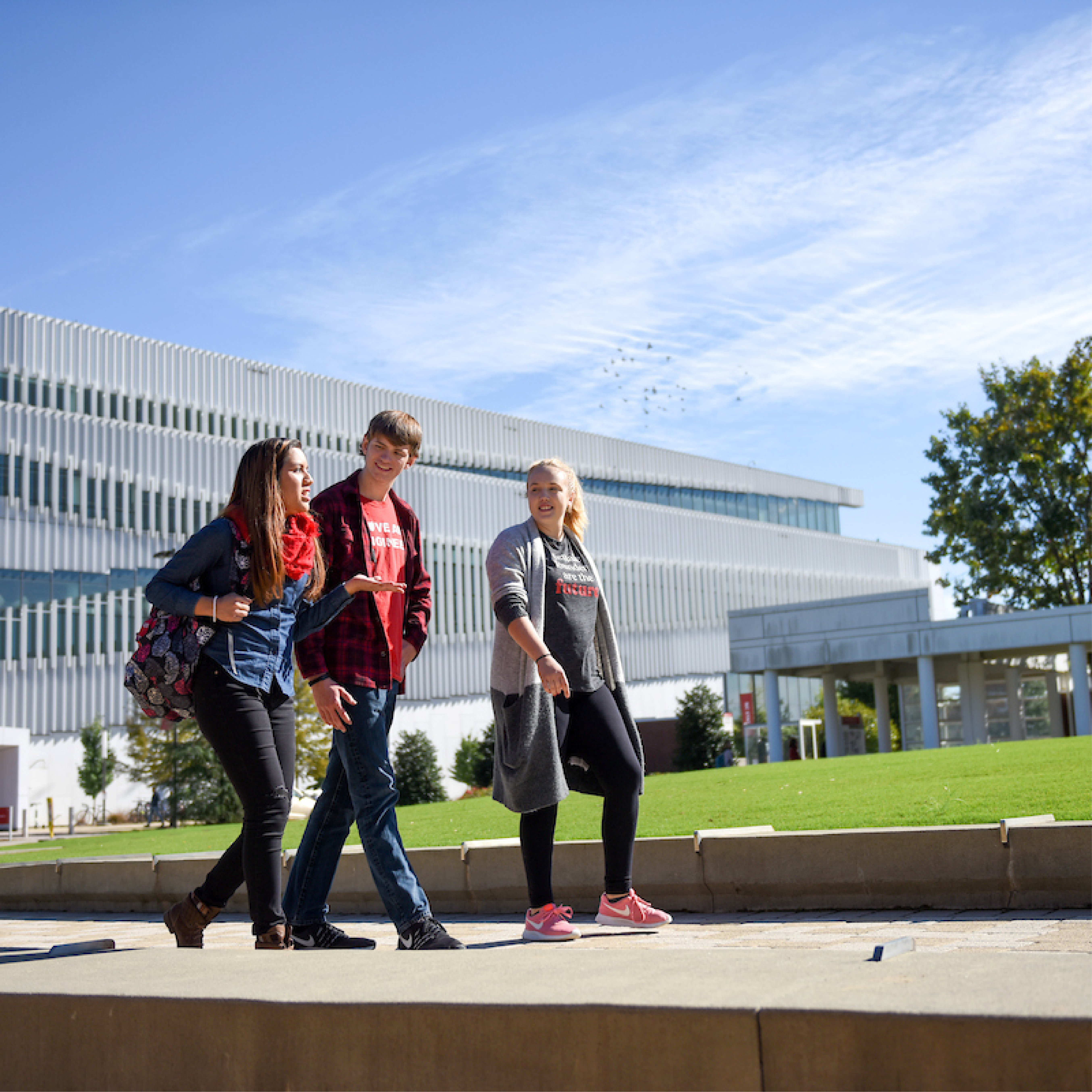 Three students walking on a sidewalk on a sunny day with a geometric building in the background