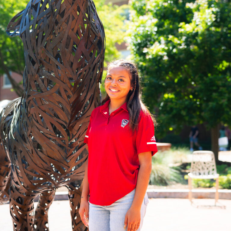 Mary Riley Locklear headshot in red shirt in front of wolf statue and green trees